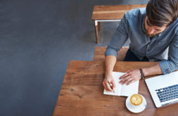 Un homme travaille à une table avec du café, un ordinateur portable, un stylo et du papier.