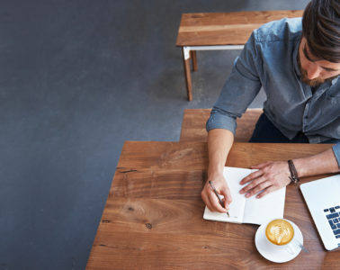 Un homme travaille à une table avec du café, un ordinateur portable, un stylo et du papier.